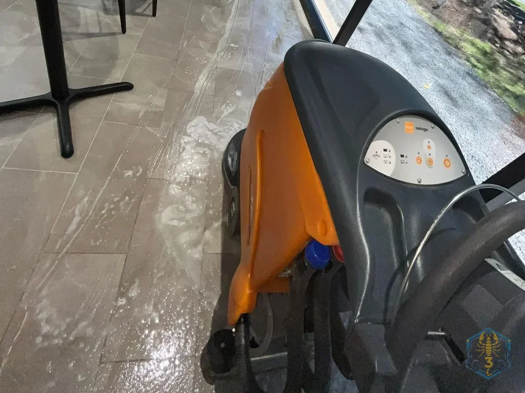 An auto-scrubber working a dining room floor at night, a wet path behind it, floor-to-ceiling windows on the right.