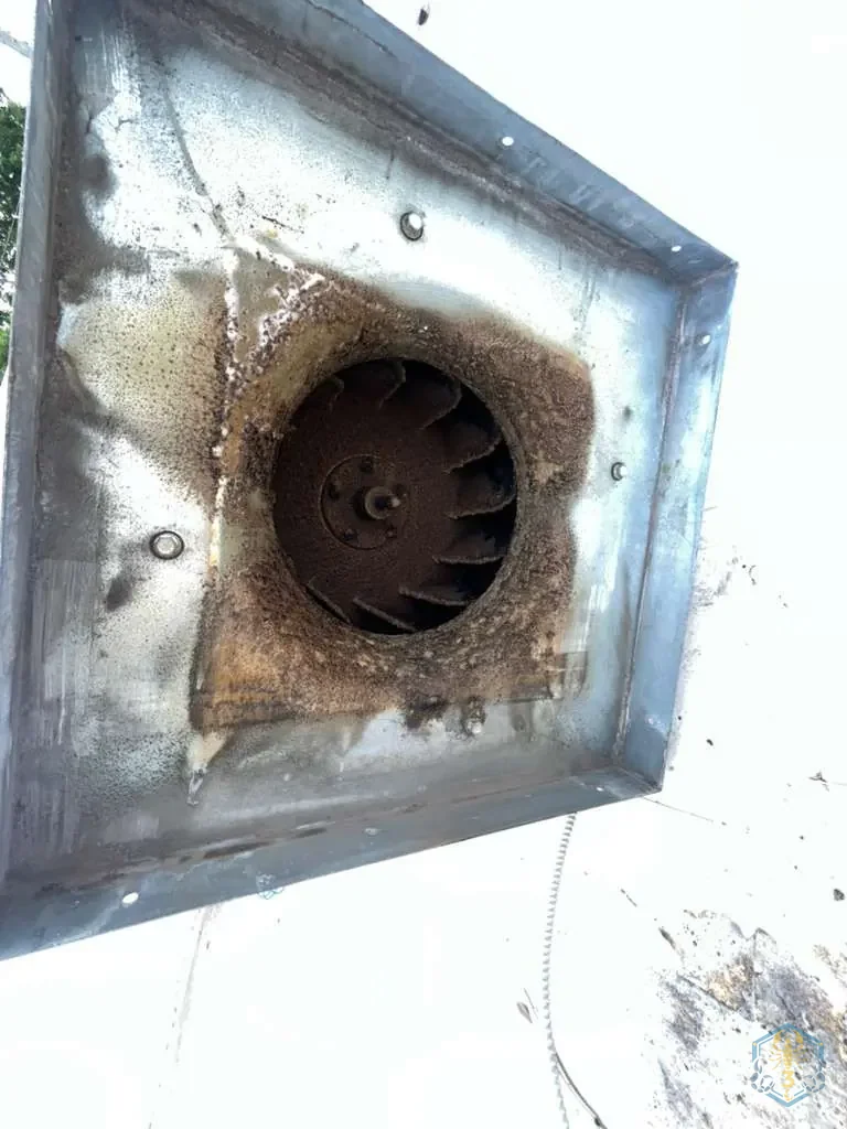Close-up of an exhaust fan housing before cleaning, with rust-brown grease coating the interior and the fan wheel.