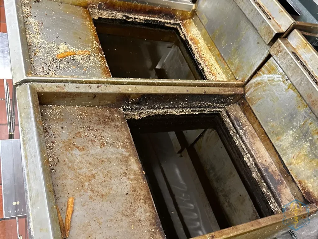 Looking down into two empty fryer vats: the decks around them are coated with grease and crumbs.